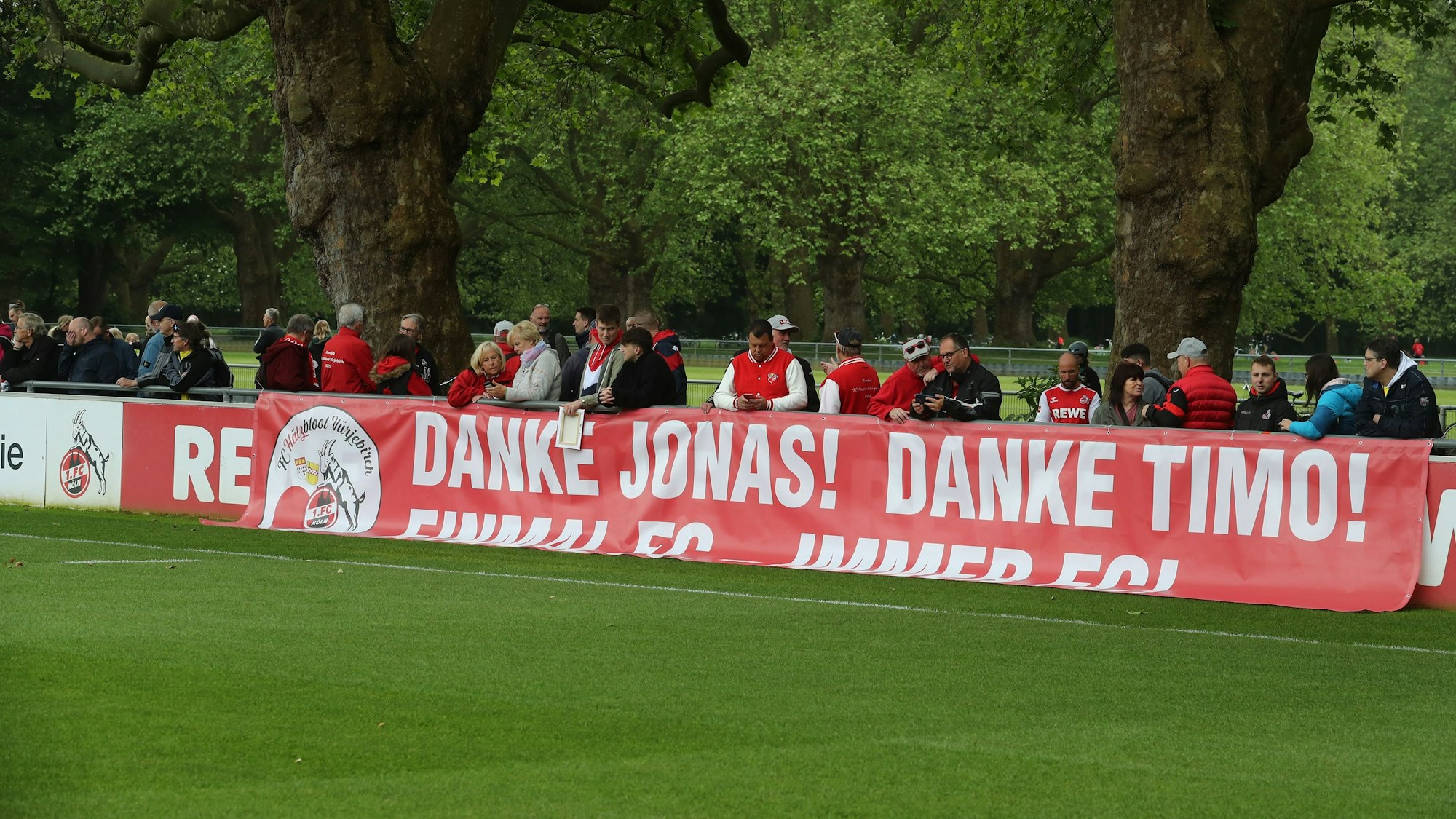 FC-Anhänger stehen beim öffentlichen Training am Geißbockheim hinter einem Fan-Plakat.