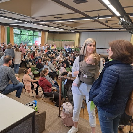 Eine Frau mit einem Kind in der Trage steht in einem Saal mit vielen Menschen.