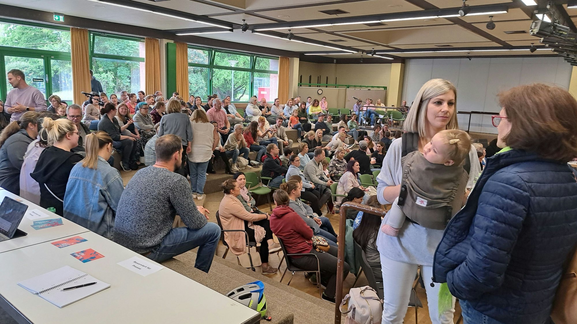 Eine Frau mit einem Kind in der Trage steht in einem Saal mit vielen Menschen.