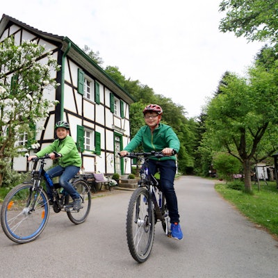 Unten am Mühlenweg ist die Radwelt noch in Ordnung, aber von hier führen nur steile Wanderwege hinauf nach Glöbusch. Zwei Kinder sitzen auf Fahrrädern.