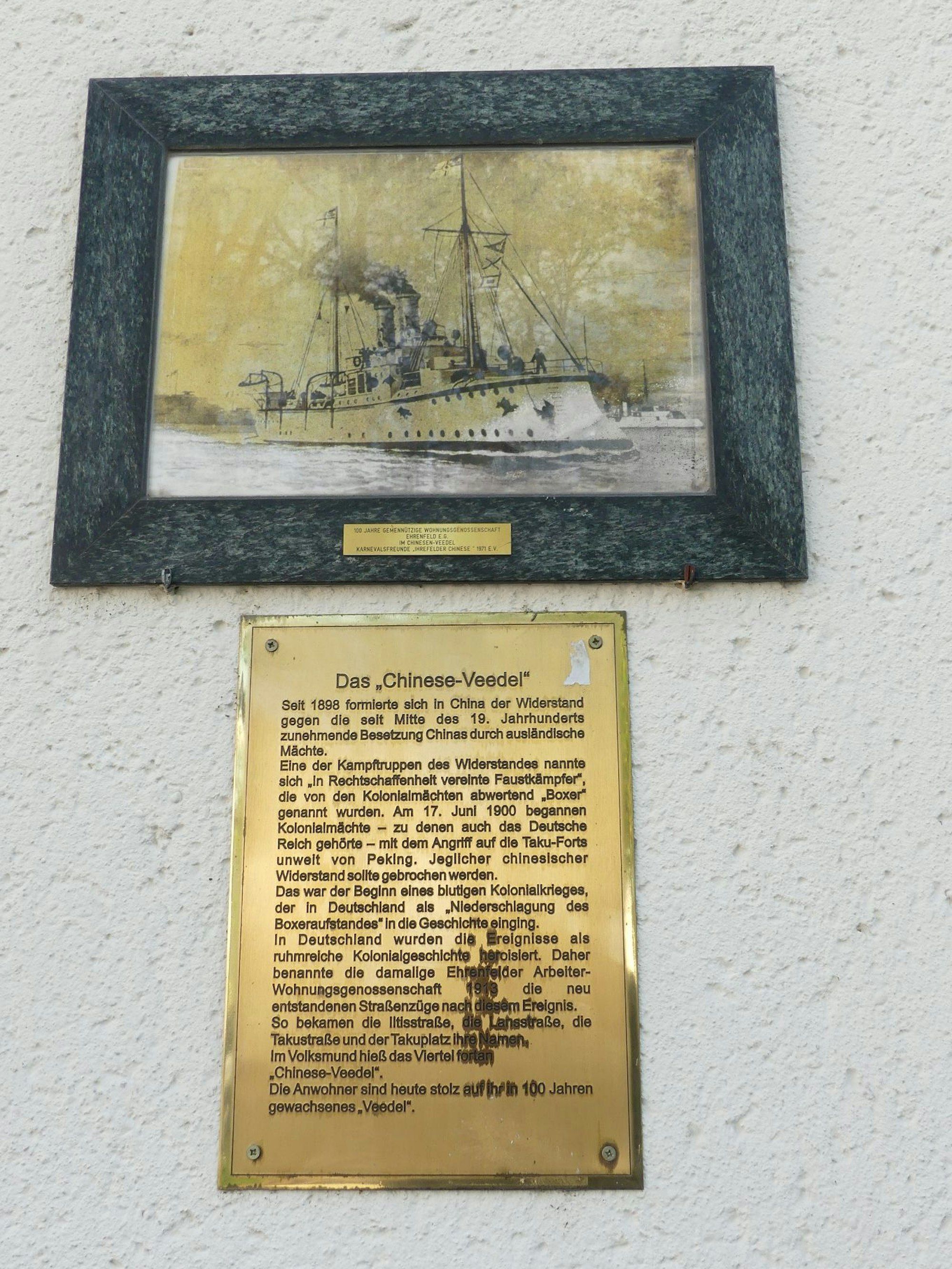 Eine Erklärtafel in Gold hängt an einer Hauswand.