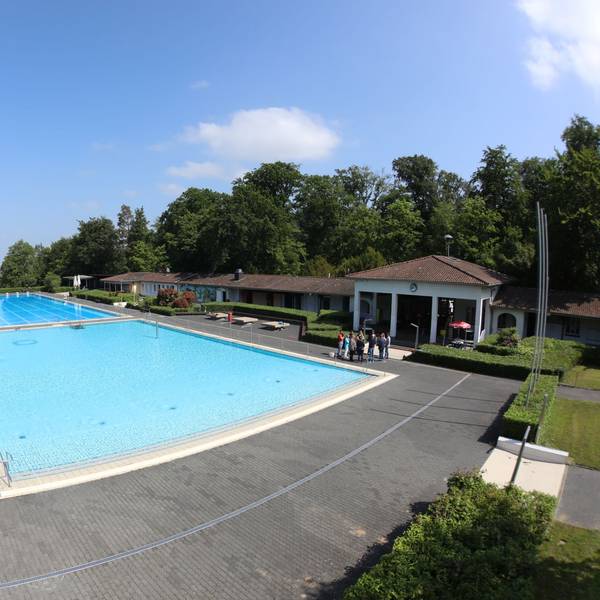 Mit Wasser gefüllt sind das Nichtschwimmer- und das 50-Meter-Schwimmerbecken. Links am Rand steht eine 65-Meter-Wasserrutsche, rechts ist das Eingangsgebäude zu sehen.