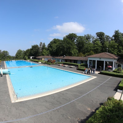 Mit Wasser gefüllt sind das Nichtschwimmer- und das 50-Meter-Schwimmerbecken. Links am Rand steht eine 65-Meter-Wasserrutsche, rechts ist das Eingangsgebäude zu sehen.