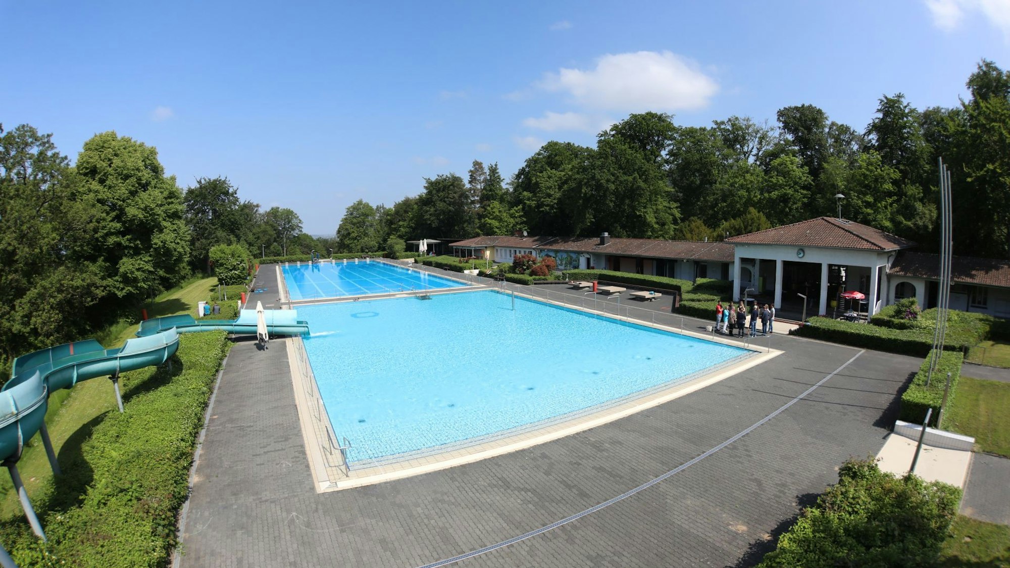 Mit Wasser gefüllt sind das Nichtschwimmer- und das 50-Meter-Schwimmerbecken. Links am Rand steht eine 65-Meter-Wasserrutsche, rechts ist das Eingangsgebäude zu sehen.