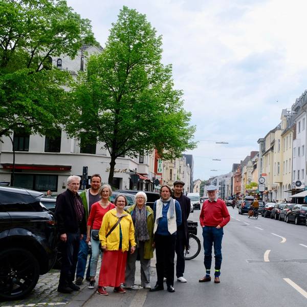 Eine Gruppe von Menschen steht am Rand einer zweispurigen Straße, im Hintergrund sind zwei Bäume zu sehen.