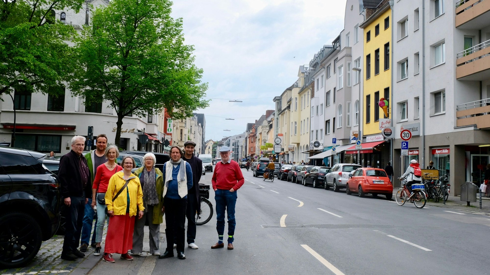 Eine Gruppe von Menschen steht am Rand einer zweispurigen Straße, im Hintergrund sind zwei Bäume zu sehen.