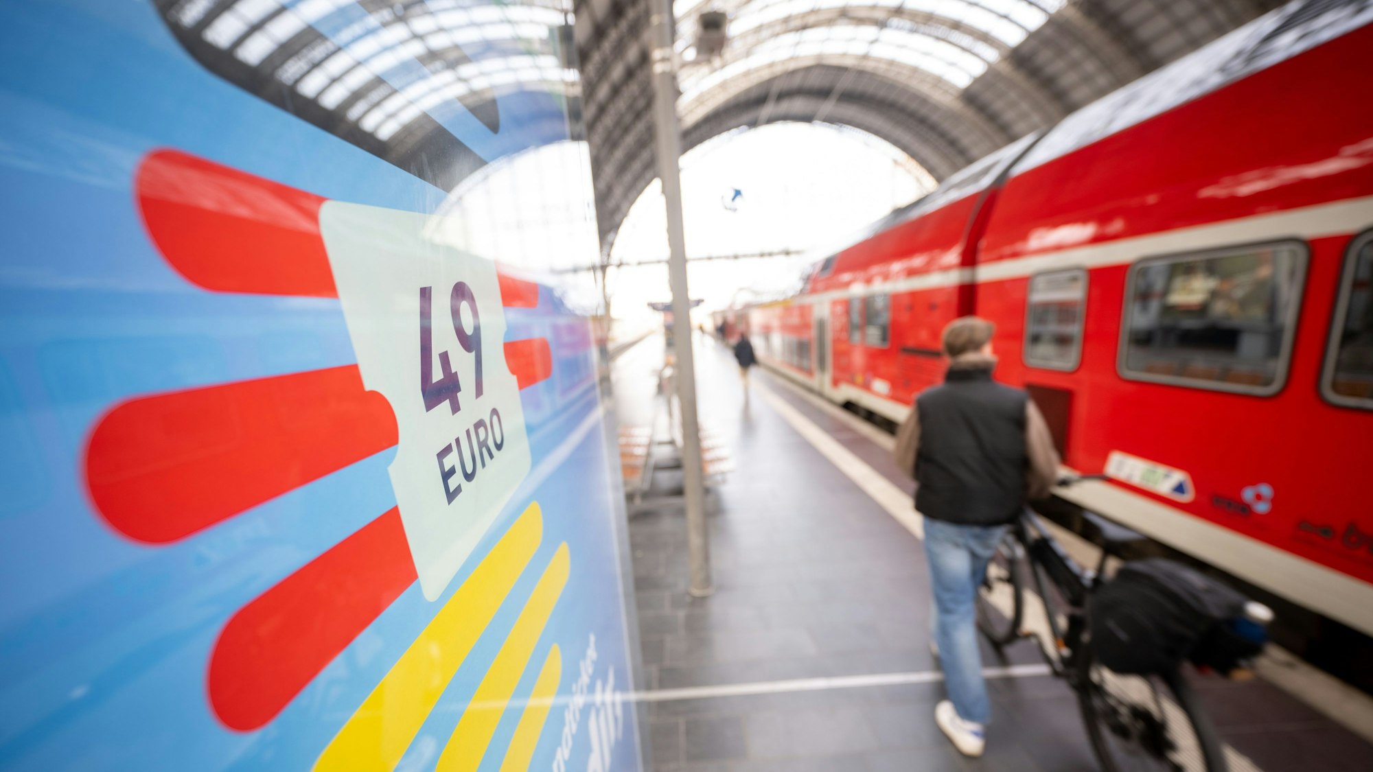 Ein Mann geht am Hauptbahnhof mit seinem Fahrrad zwischen der Werbung für das Deutschlandticket und einer Regionalbahn vorbei. Seit einem Monat gilt bundesweit das 49 Euro teure Deutschlandticket im öffentlichen Personennahverkehr.