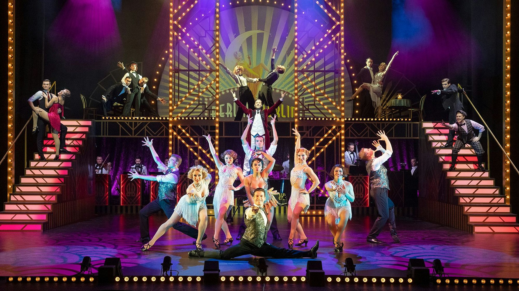Die Schauspieler und Schauspielerinnen von „Berlin Berlin“ stehen zusammen auf einer bunt erleuchteten Bühne.
