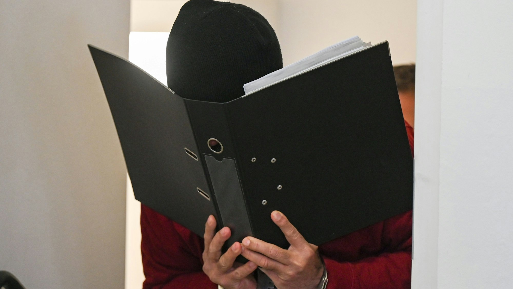 Der Angeklagte, der in Handschellen ist und sein Gesicht hinter einem Aktenordner versteckt, wird zum Prozessauftakt in den Gerichtssaal des Landgerichts geführt.