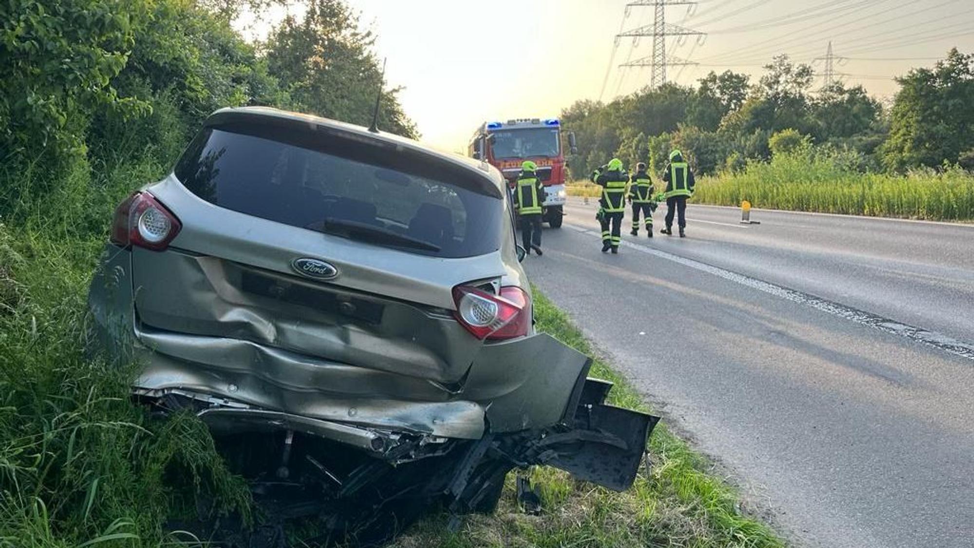 Ein graues Auto steht auf einem Grünstreifen, der hintere Teil des Wagens ist verbeult. Im Hintergrund sind Feuerwehrleute zu sehen.
