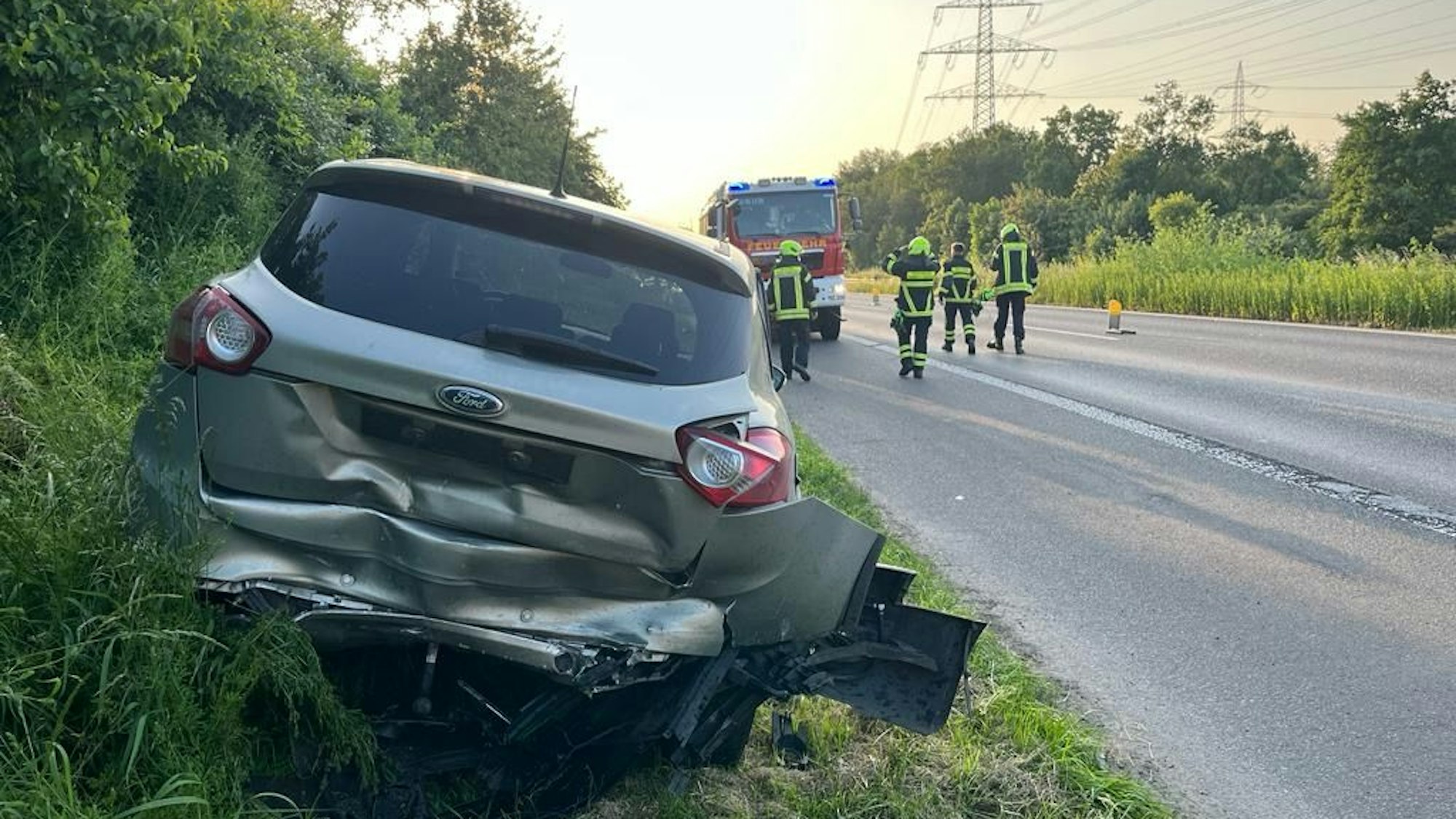Ein graues Auto steht auf einem Grünstreifen, der hintere Teil des Wagens ist verbeult. Im Hintergrund sind Feuerwehrleute zu sehen.