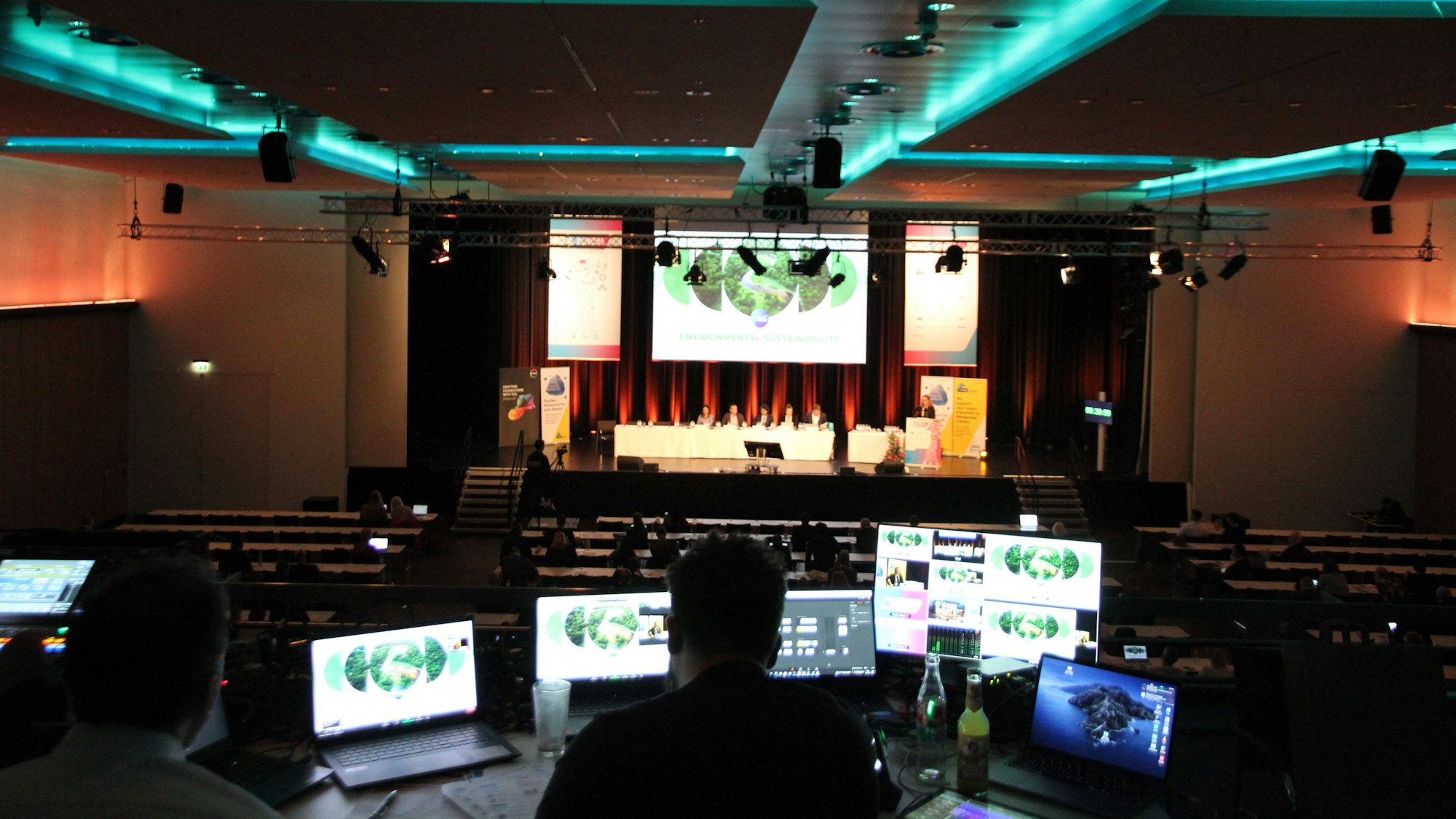 Zwei Männer an Computermonitoren auf der Empore eines großen Veranstaltungssaals mit einer Podiumsdiskussion auf der Bühne.
