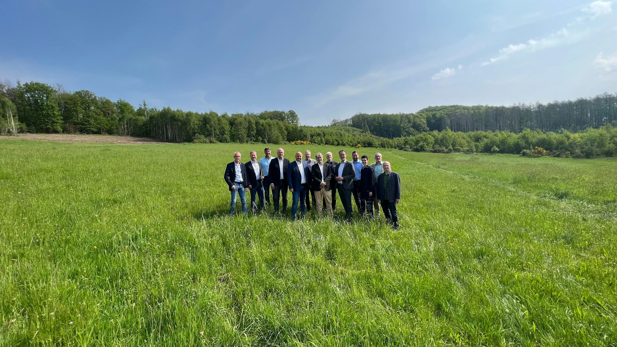 Eine große, grüne Wiese mit Bäumen im Hintergrund und strahlend blauem Himmel. In der Mitte steht eine Gruppe aus 13 Personen.