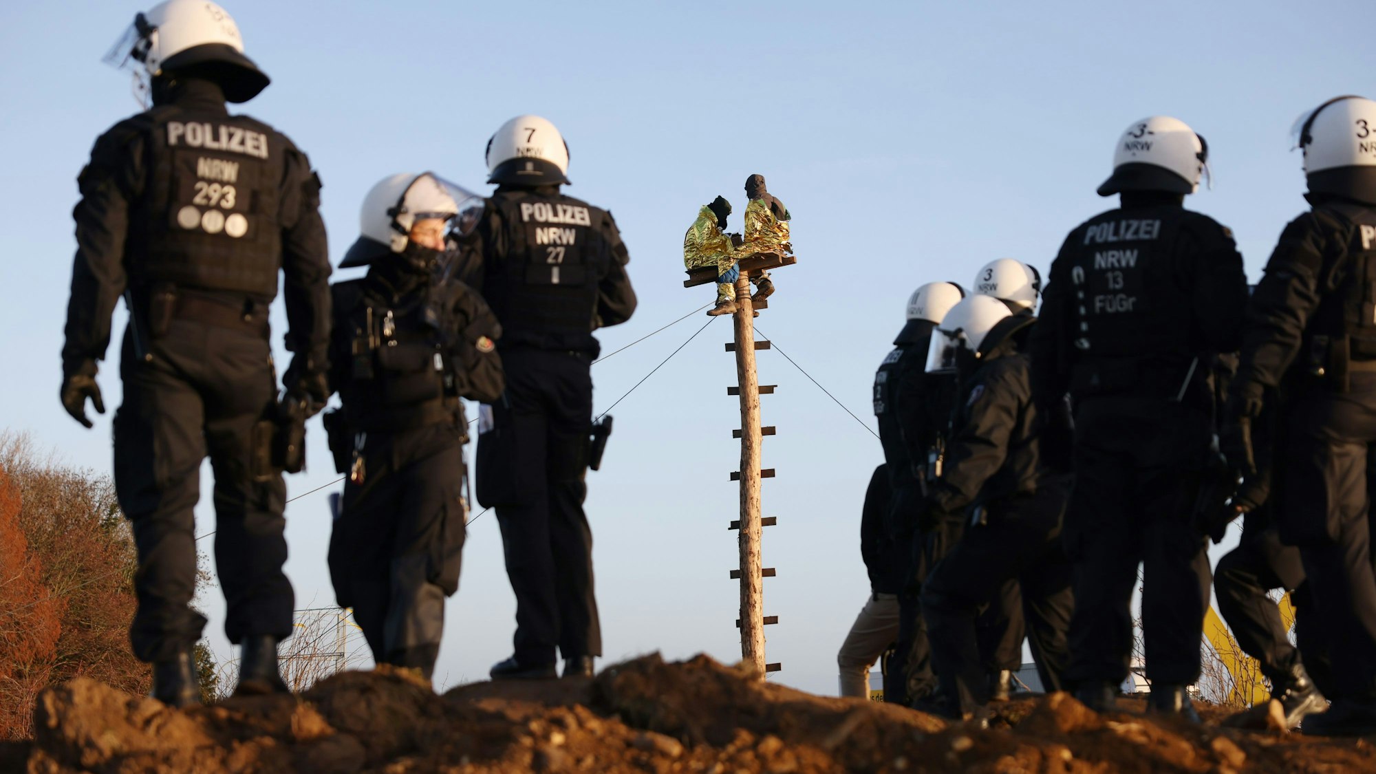 Klimaschutzaktivisten sitzen auf einem Holzpfahl am Rand der Ortschaft Lützerath. Am Boden stehen Polizisten einer Hundertschaft.