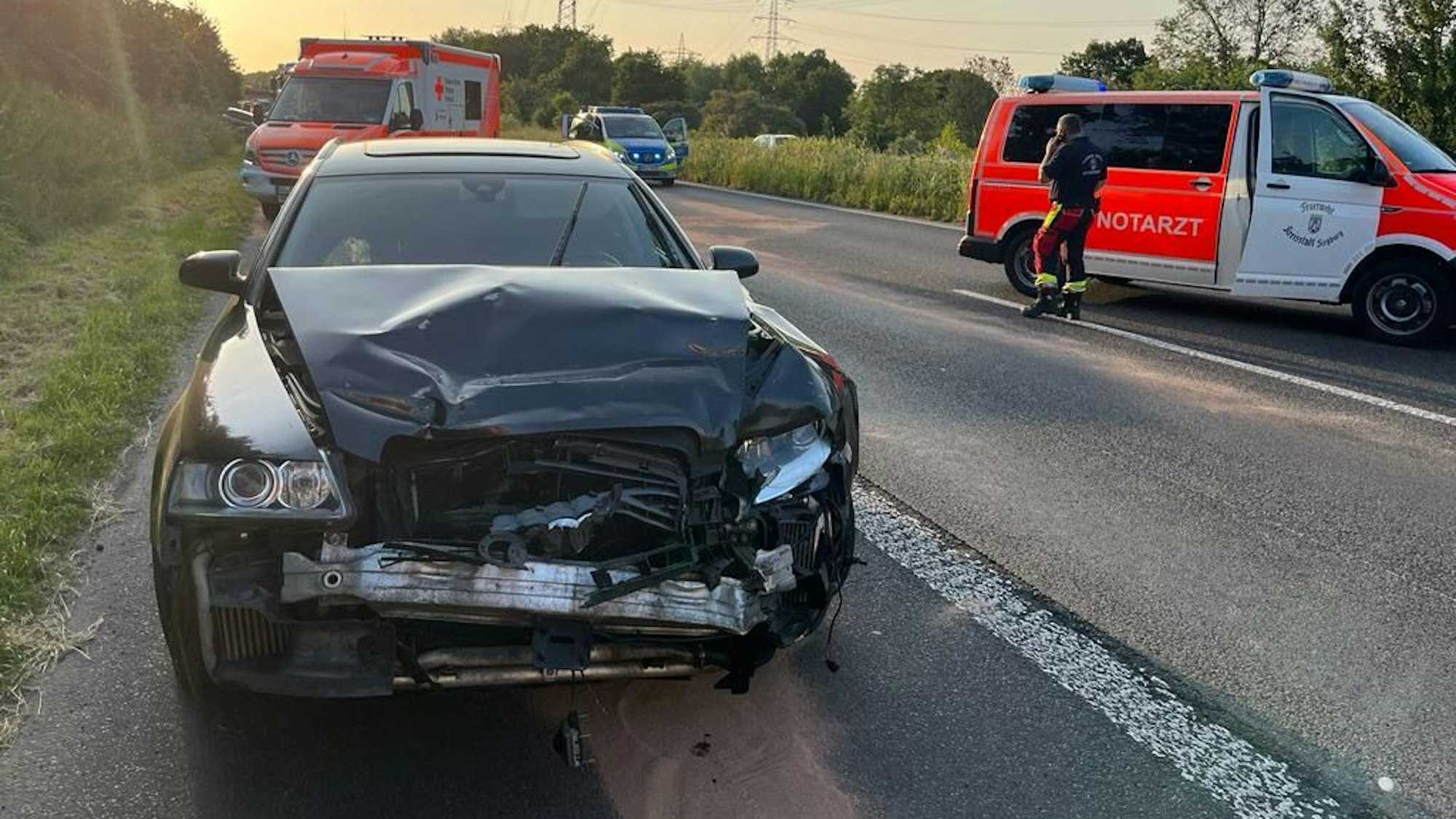 Ein schwarzes Auto steht auf dem Standstreifen, der vordere Teil des Wagens ist verbeult. Im Hintergrund sind Autos von Notarzt, Polizei und Feuerwehr zu sehen.
