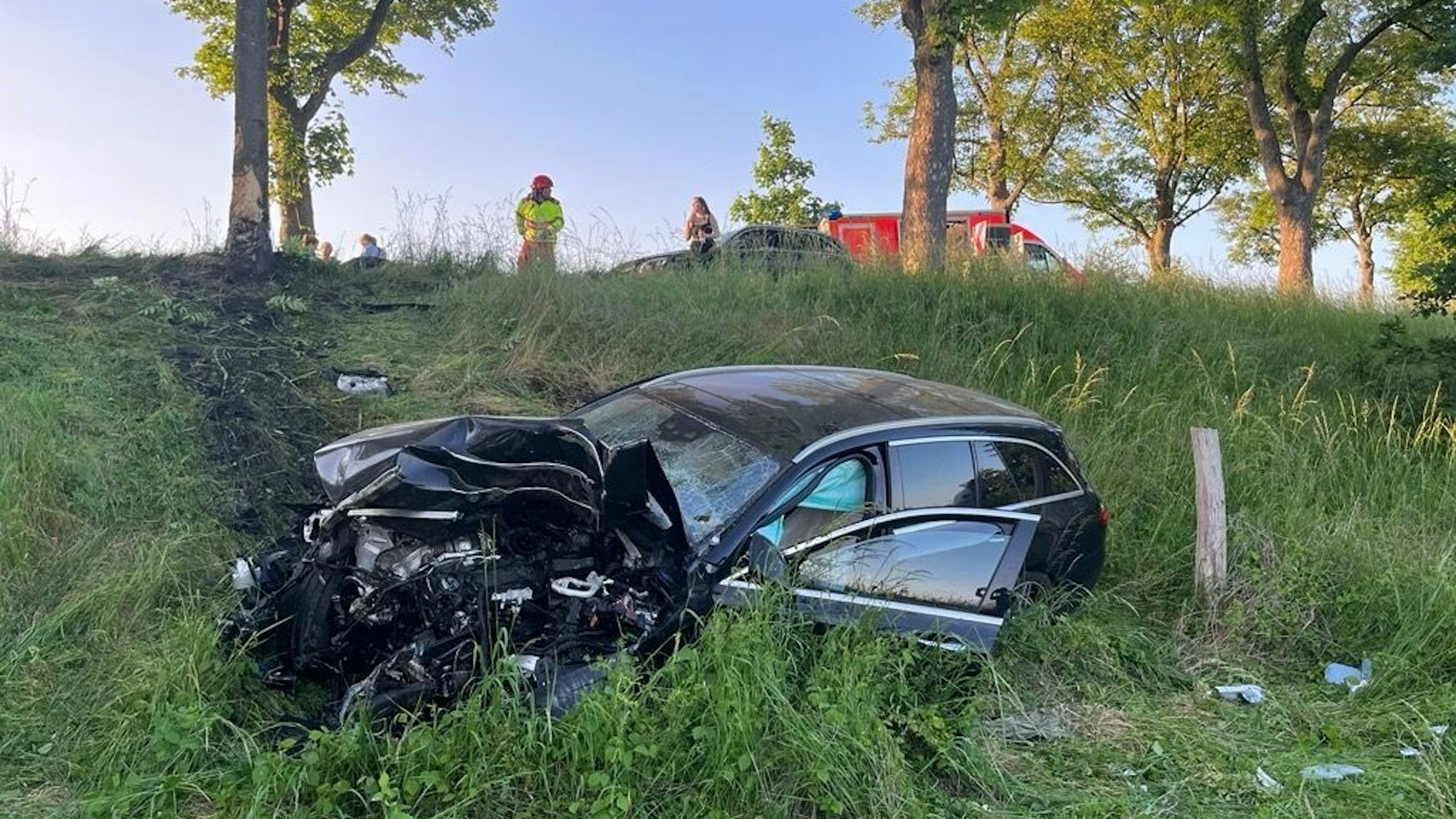 Ein komplett kaputtes Auto liegt auf einer Wiese. Reifenspuren sind deutlich sichtbar, im Hintergrund steht ein Feuerwehrmann.