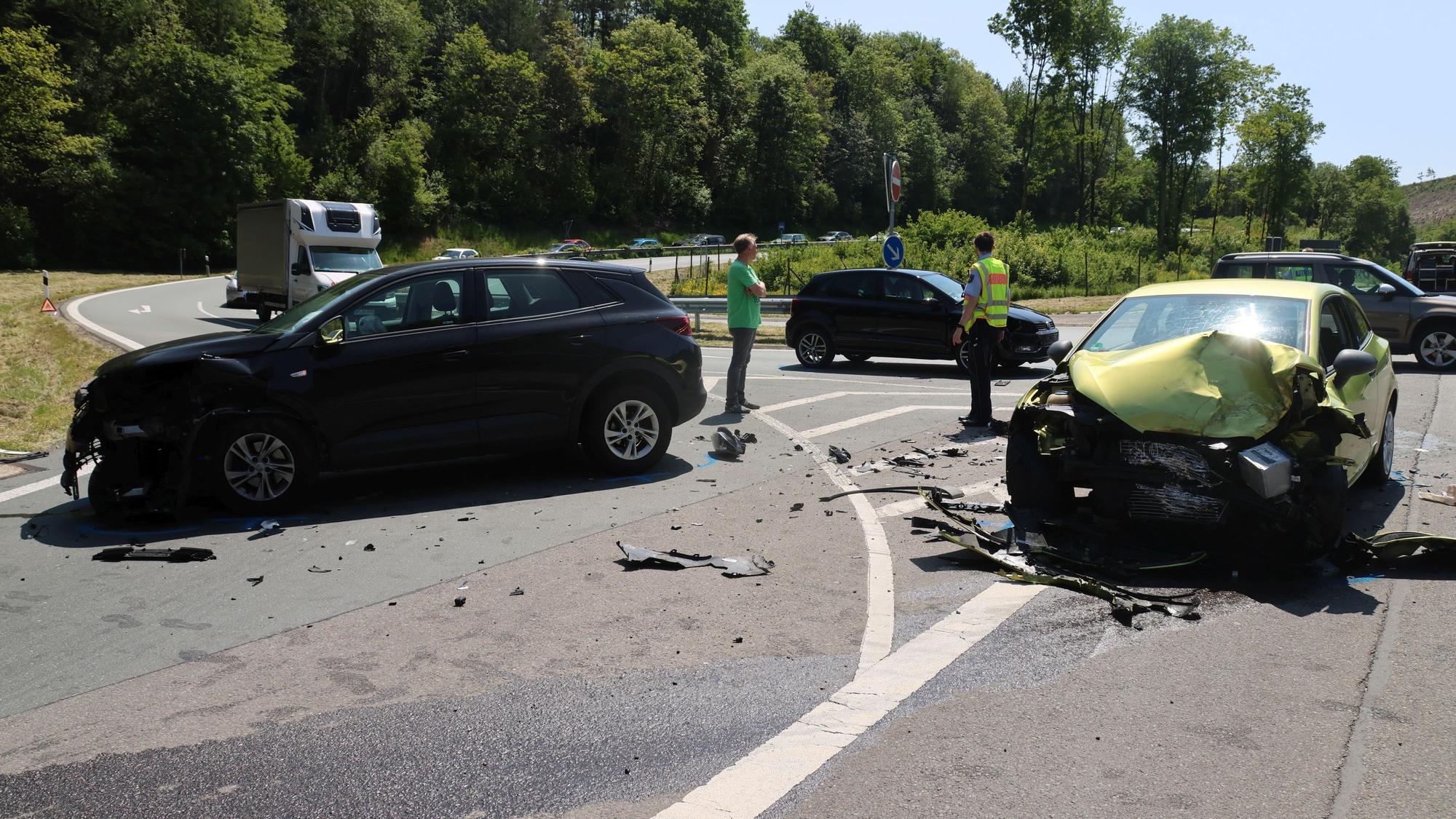 Drei Autos stehen schwer beschädigt auf der Straße. Zwischen Ihnen eine Person in grünem T-Shirt und ein Polizist.