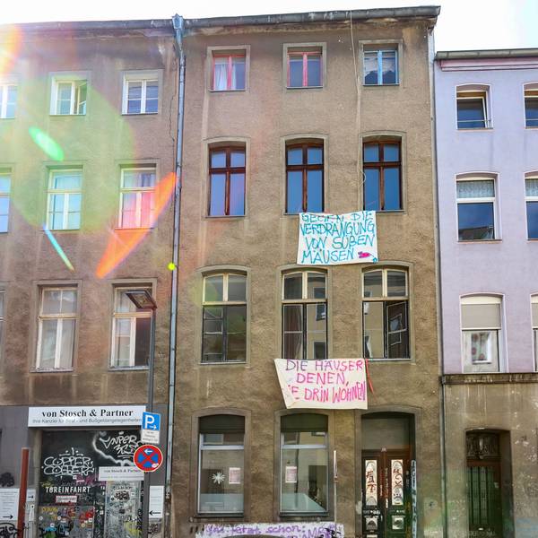Eine Häuserzeile mit vier mehrgeschossigen Häusern ist zu sehen. Vor der Front des braunen Hauses hängen Banner mit Slogans, etwa Die Häuser denen, die drin wohnen.
