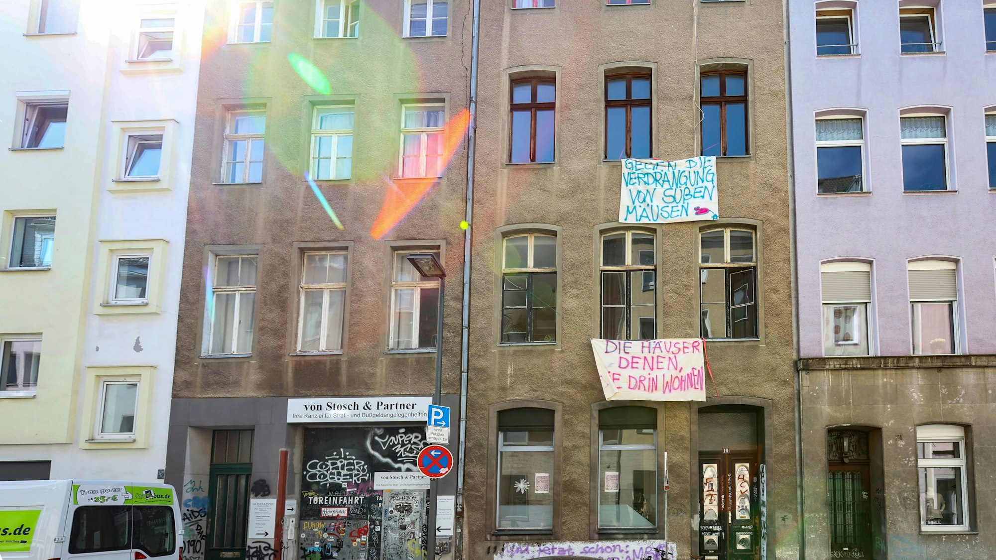 Eine Häuserzeile mit vier mehrgeschossigen Häusern ist zu sehen. Vor der Front des braunen Hauses hängen Banner mit Slogans, etwa Die Häuser denen, die drin wohnen.