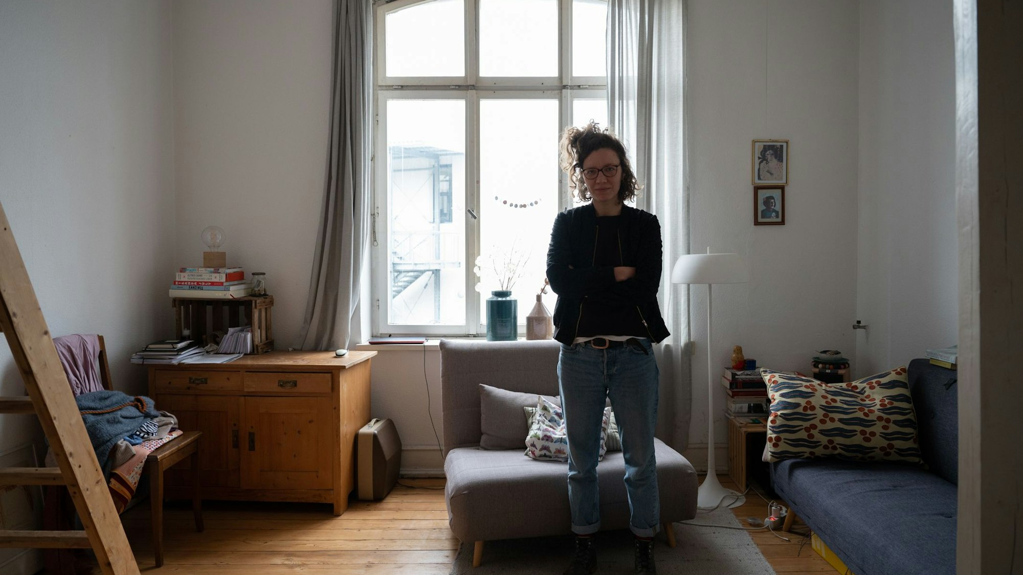 Eine junge Frau steht mit verschränkten Armen vor einem Sessel in einem Zimmer mit Holzboden und hoher Decke.