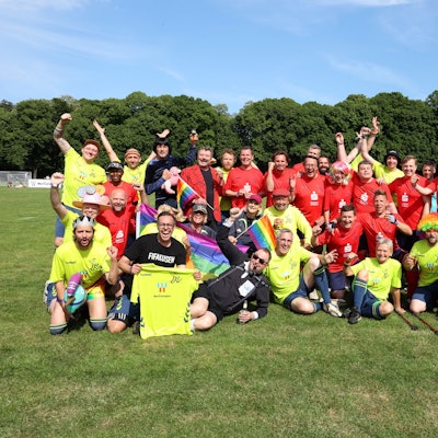 Beim Promi-Spaßkick im Rahmen des Come-Together-Cups vor dem FC-Stadion am Sonntag (28. Mai 2023) mischten einige bekannte Gesichter mit. Personen in gelben und roten Trikots posieren in zwei Reihen. Einige Personen schwenken dabei Regenbogenfahnen.