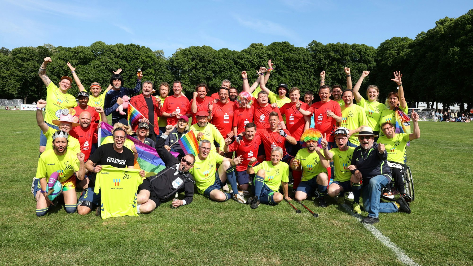Beim Promi-Spaßkick im Rahmen des Come-Together-Cups vor dem FC-Stadion am Sonntag (28. Mai 2023) mischten einige bekannte Gesichter mit. Personen in gelben und roten Trikots posieren in zwei Reihen. Einige Personen schwenken dabei Regenbogenfahnen.