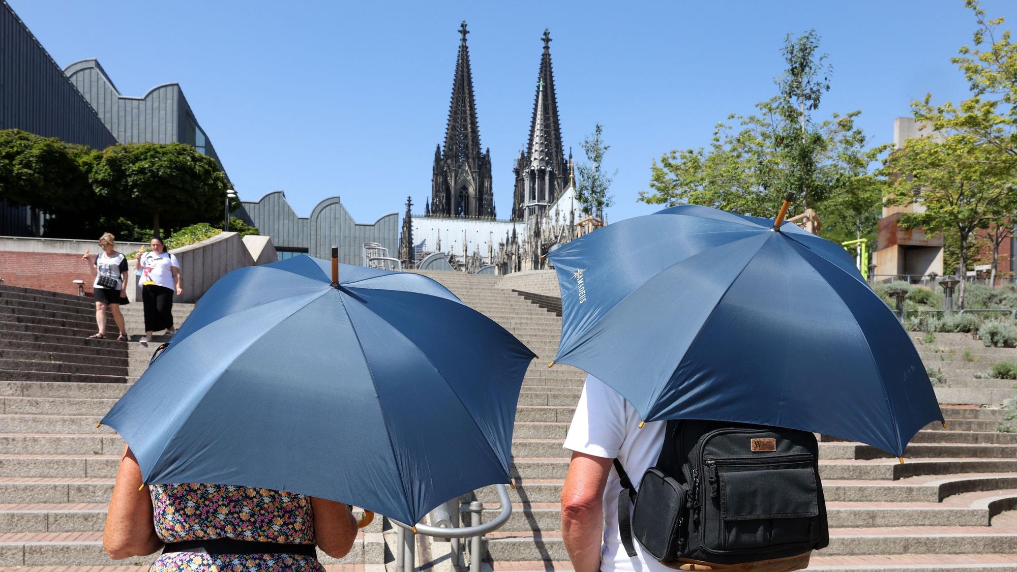 Zwei Touristen laufen mit Regenschirmen, die als Sonnenschirme genutzt werden, auf den Dom zu.