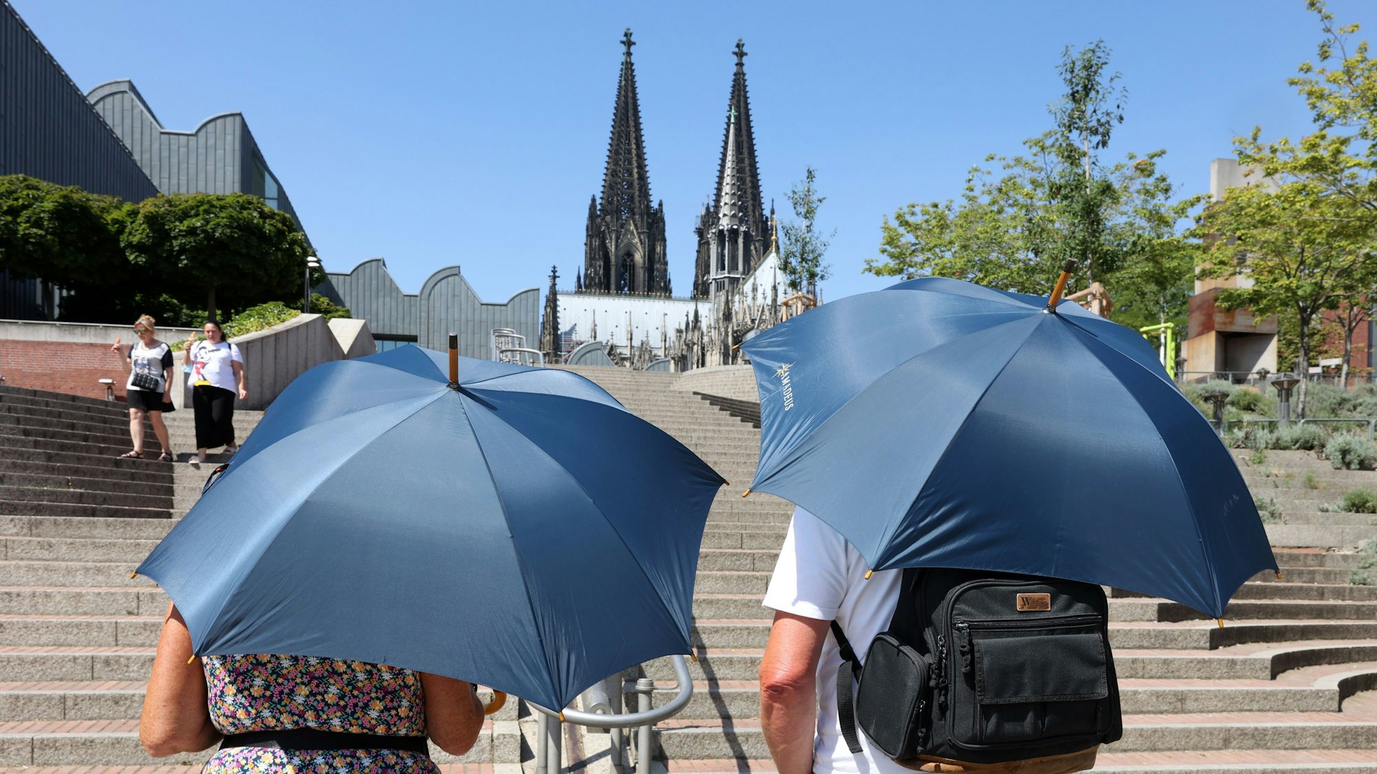 Zwei Touristen laufen mit Regenschirmen, die als Sonnenschirme genutzt werden, auf den Dom zu.