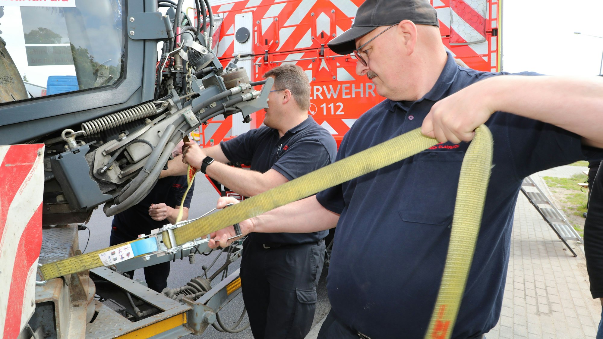 Zwei Männer befestigen einen Spanngurt an einem Tieflader und einem Fahrzeug darauf.