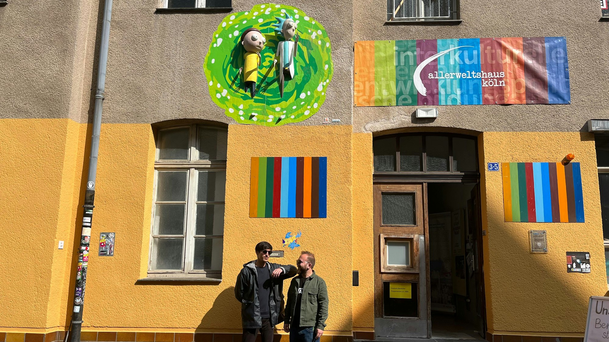 Yaşar Fidan (l.) und Lemi Karaca von Coach e.V. stehen vor ihrem Kunstwerk am Allerweltshaus in Ehrenfeld. Die 3D-Installation zeigt Rick und Morty aus der gleichnamigen Serie, wie sie aus einem Portal kommen.