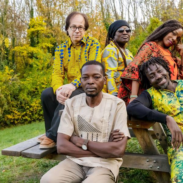 Die Band trägt traiditonelle, größtenteils bunte Outfits. Sie sitzen an einer Bank mit Tisch im Grünen.
