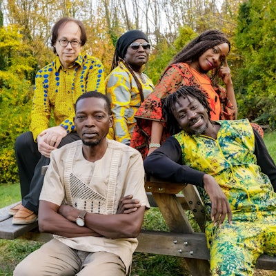 Die Band trägt traiditonelle, größtenteils bunte Outfits. Sie sitzen an einer Bank mit Tisch im Grünen.