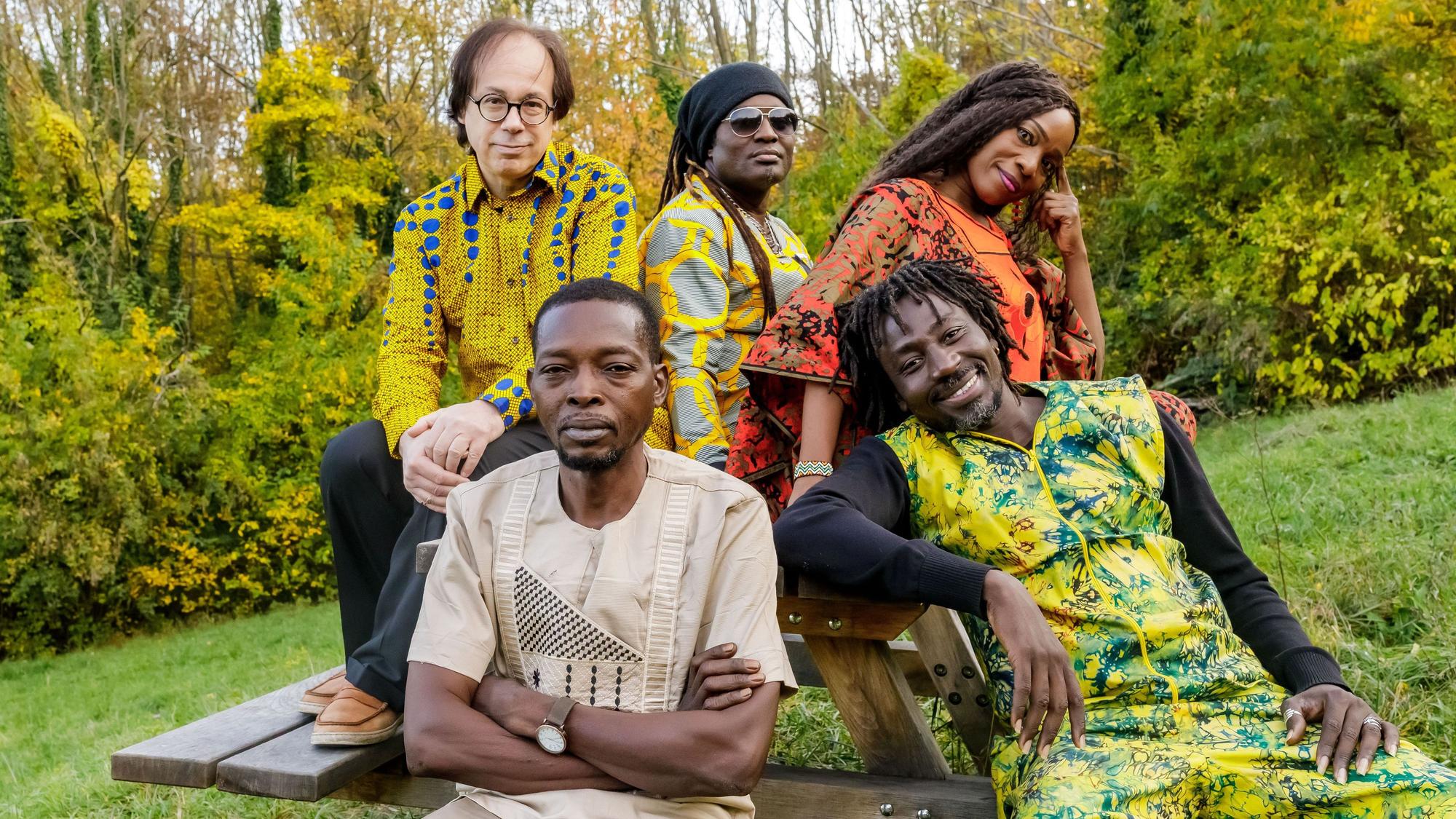 Die Band trägt traiditonelle, größtenteils bunte Outfits. Sie sitzen an einer Bank mit Tisch im Grünen.