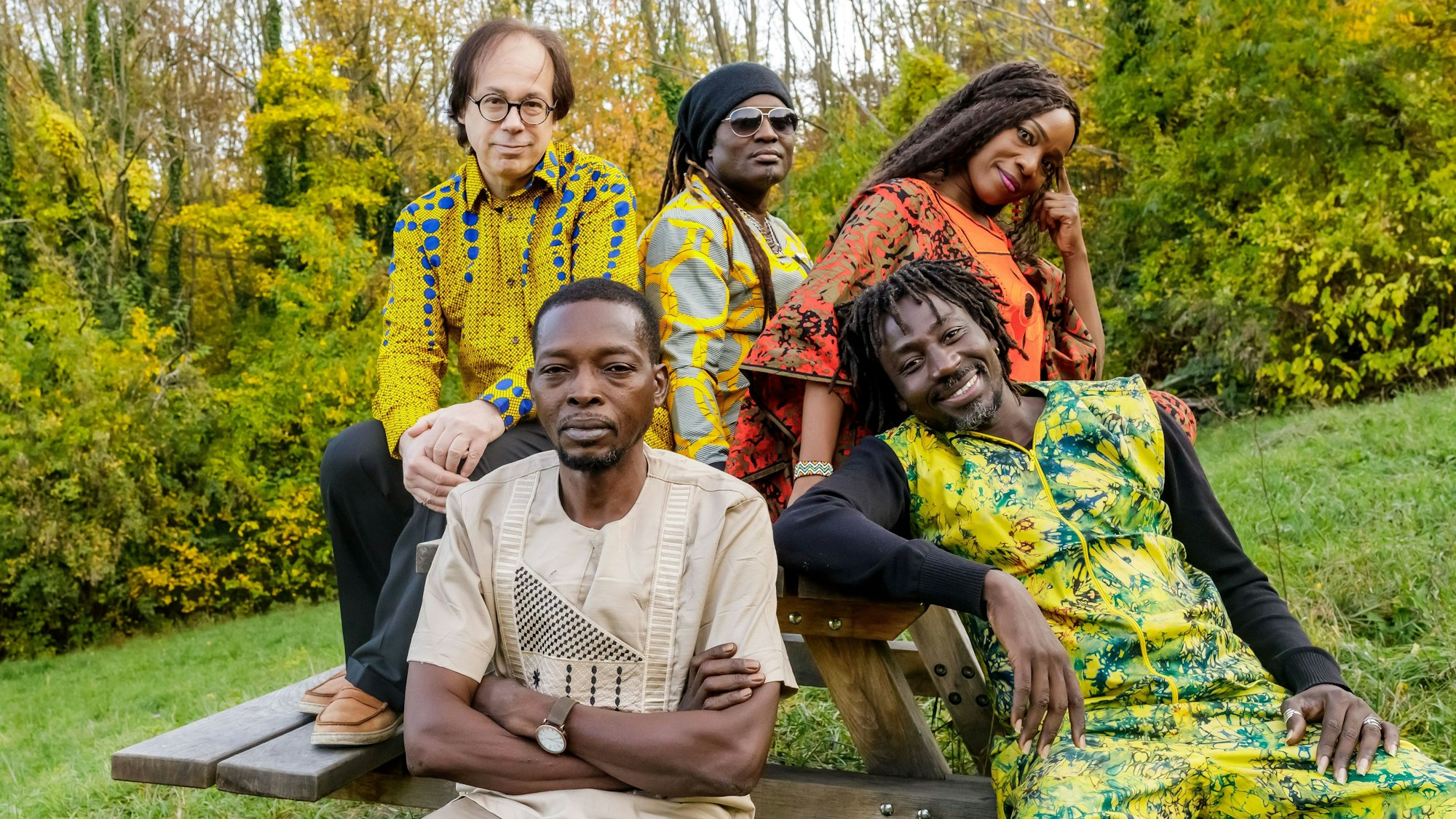 Die Band trägt traiditonelle, größtenteils bunte Outfits. Sie sitzen an einer Bank mit Tisch im Grünen.