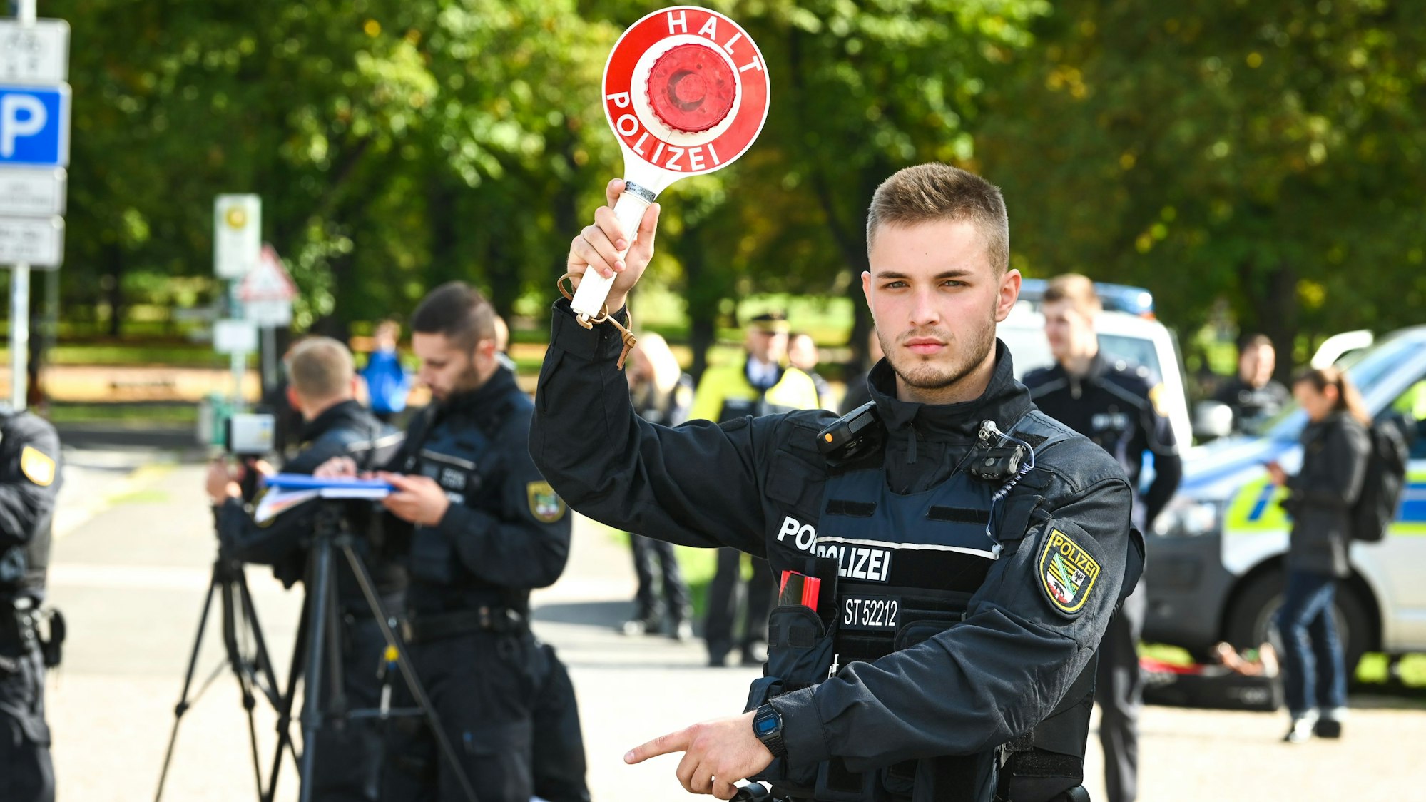 Ein Beamter des Zentralen Einsatzdienstes der Polizei hält eine Kelle mit der Aufschrift „Halt Polizei“ hoch, um Autos anzuhalten. Im Bildhintergrund befinden sich Beamte mit Geschwindigkeitsmessgeräten und weitere Beamte vor Einsatzfahrzeugen.