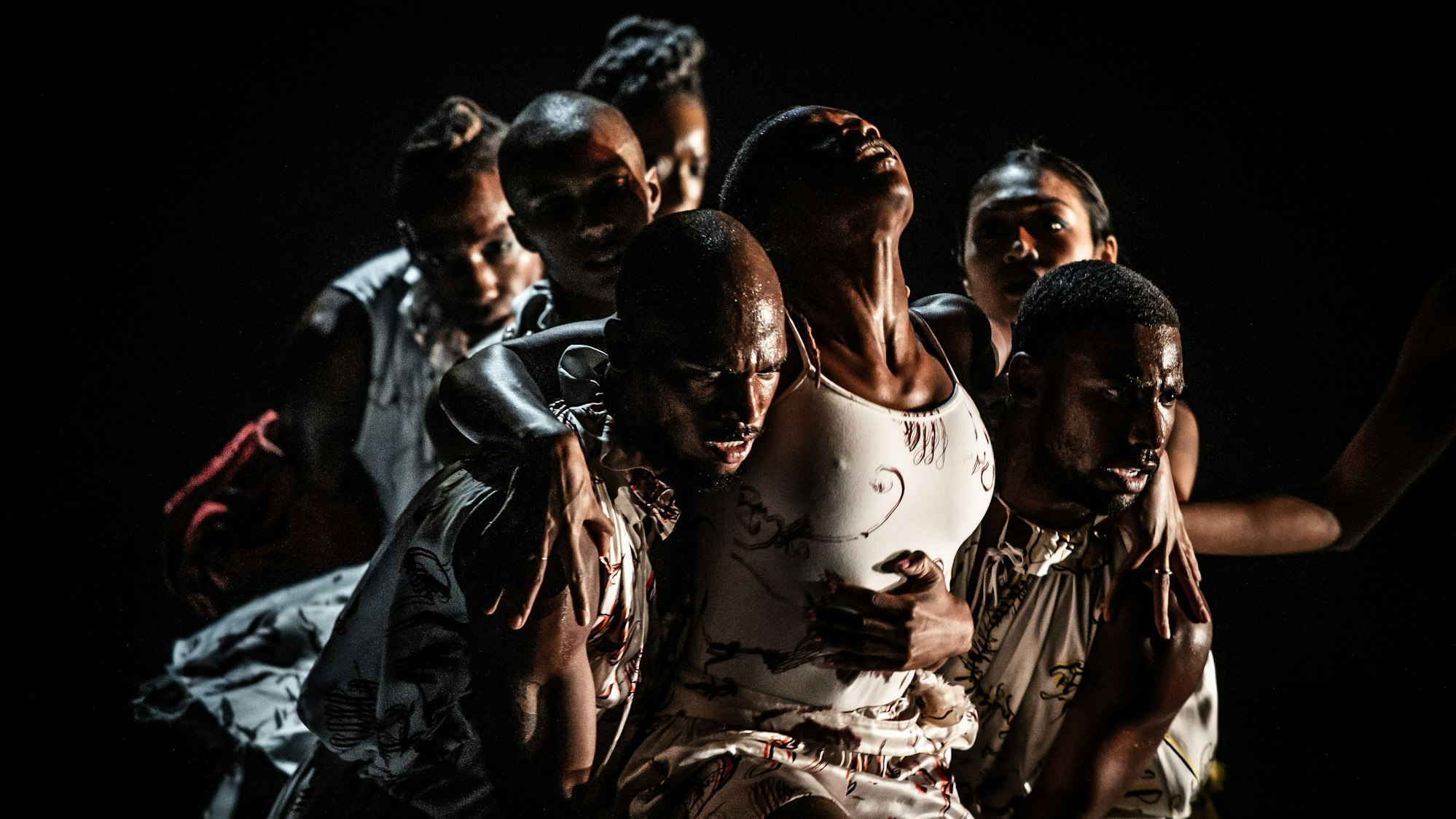 Zwei Tänzer tragen eine Tänzerin, die zum Himmel schaut. Im Hintergrund weitere Tänzer. Sie schauen angespannt.