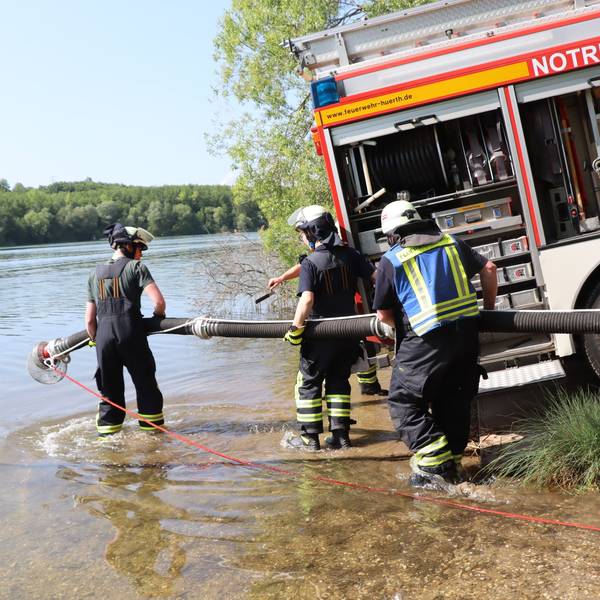 Ein Feuerwehrauto steht am Rand des Sees, Feuerwehrleute verlegen einen Schlauch ins Wasser.