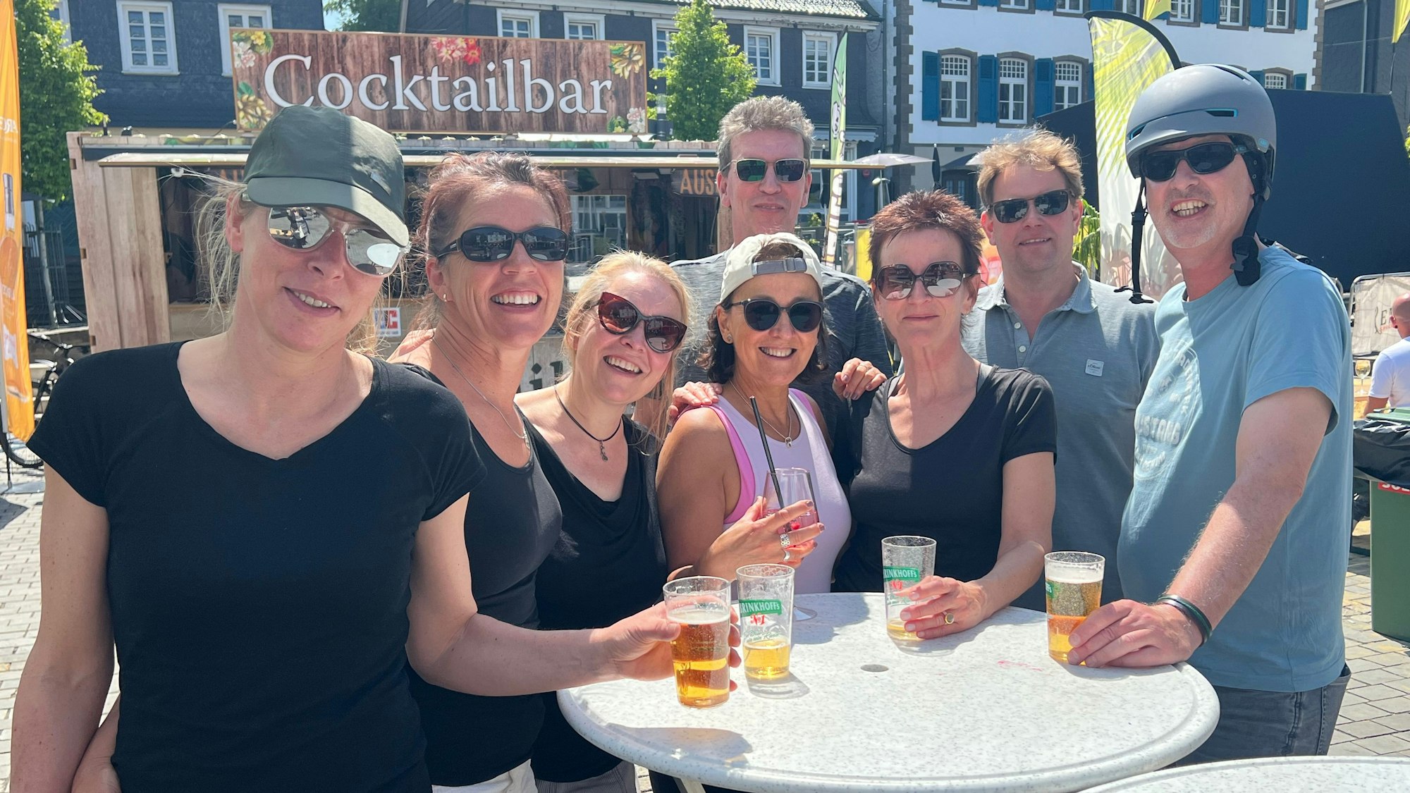Eine Gruppe von acht Personen vor einem Stehtisch, einige von ihnen haben einen Becher vor sich. Die Sonne scheint, im Hintergrund die Cocktailbar.