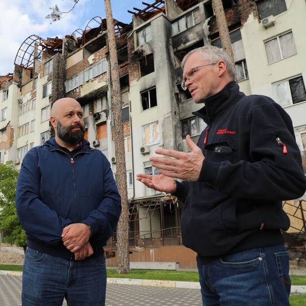Drei MEnschen stehen sprechend vor einem zerbombten und ausgebrannten Wohn- und Geschäftshaus bei Butscha.