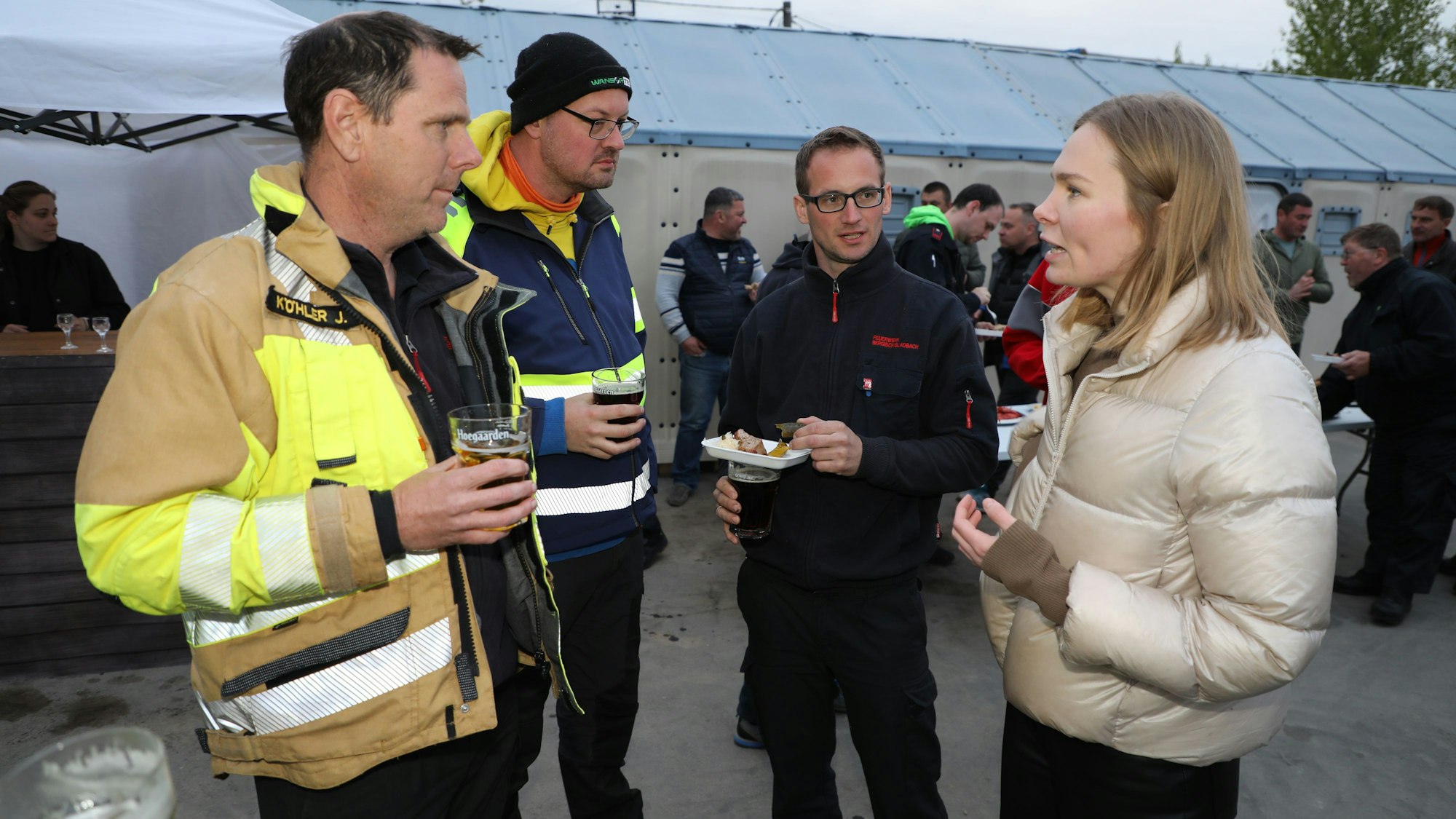 Dei Männer und eine Frau stehen mit Getränken und Gegrillten in den Händen auf einem Platz eines Bauhofgeländes.