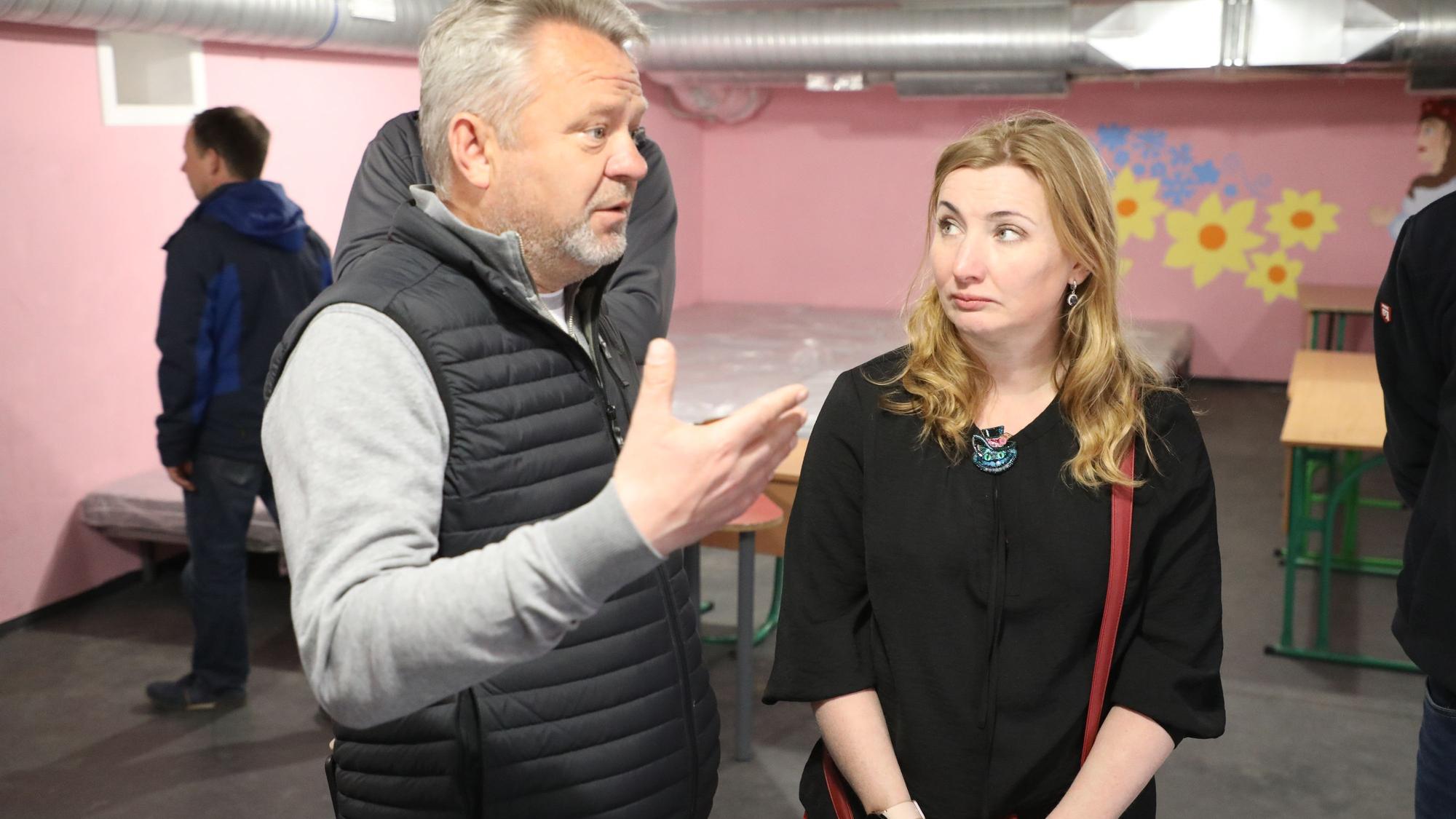 Ein Mann und eine Frau stehen in einem Bunker, an dessen Wände Blumen in Geld und Blau gemalt sind. Im Hintergrund stehen Betten und Schultische.