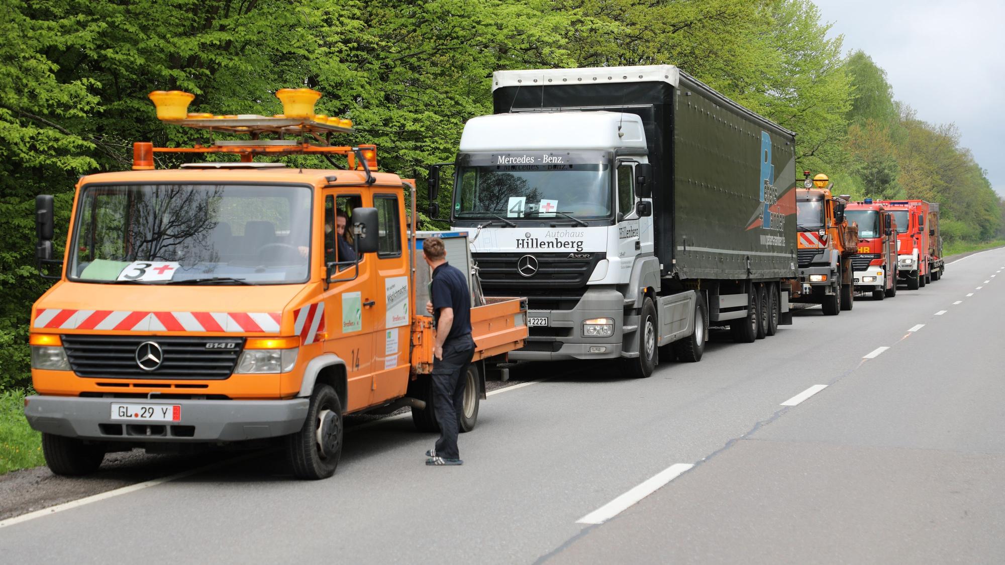 Autos und Lkw, darunter Straßenmeisterei- und Feuerwehrfahrzeuge, stehen am Straßenrand.