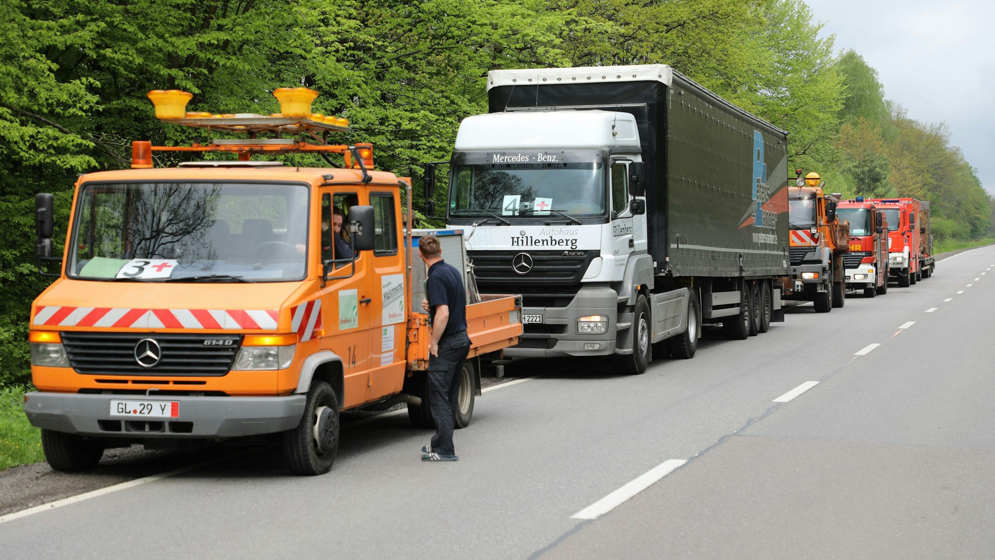 Autos und Lkw, darunter Straßenmeisterei- und Feuerwehrfahrzeuge, stehen am Straßenrand.