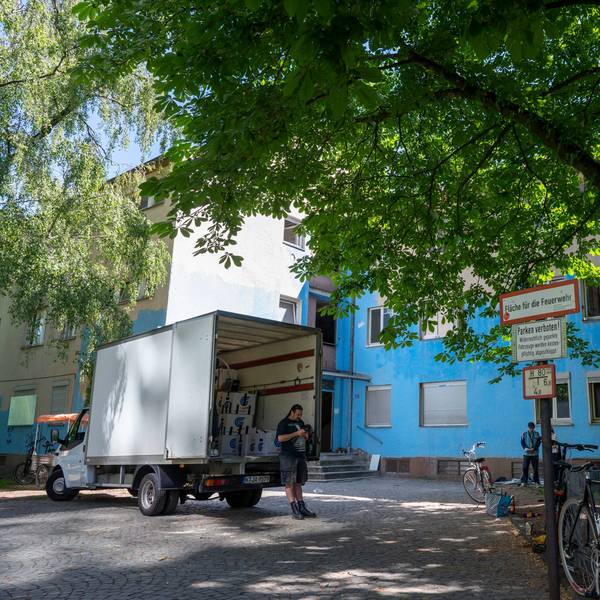 Weißer Lieferwagen vor einem blau angemalten, heruntergekommen aussehenden Gebäude.