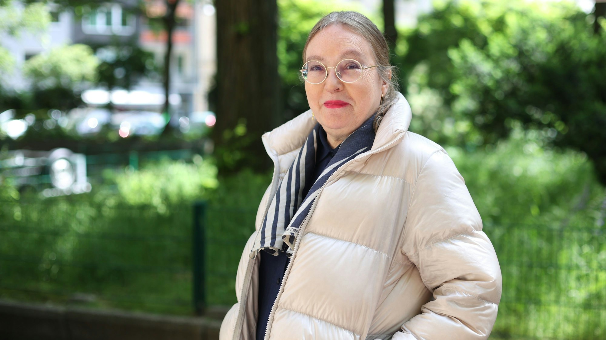 Frau Voggenreiter steht auf dem Brüsseler Platz, im Hintergrund viel grün. Sie trägt eine weiße Steppjacke und eine runde Brille.