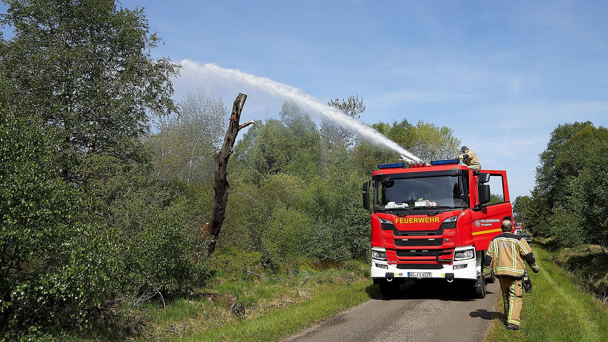 Ein Löschfahrzeug spritzt Wasser in die Büsche, ein Feuerwehrmann steht vor dem Auto.