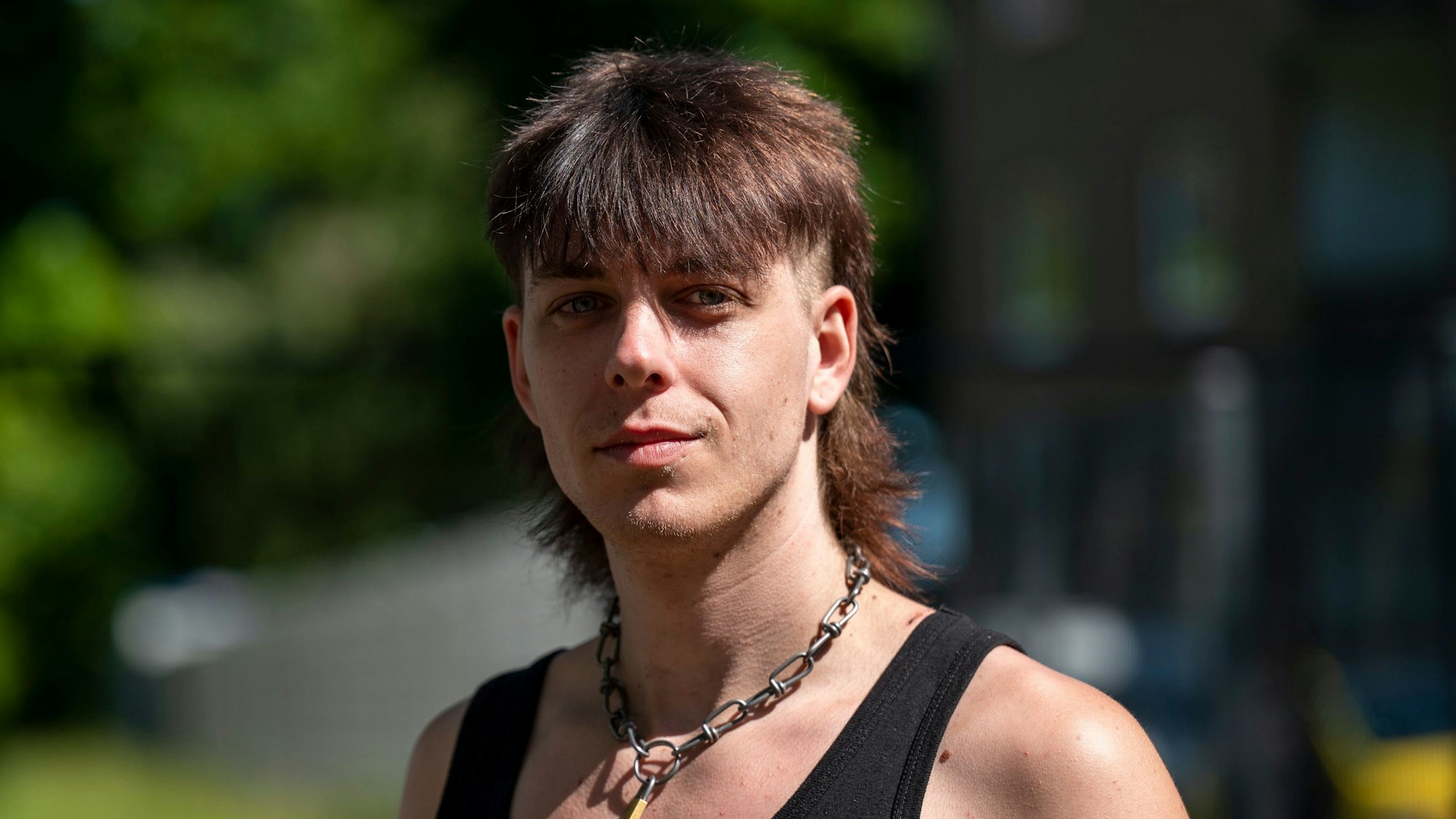 Junger Mann in einem schwarzen Tanktop mit einem Fokuhila und einem Kettenschloss um dem Hals