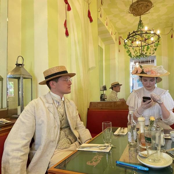 Ein Mann mit Hut und hellem Anzug mit Weste und Krawatte und eine Frau mit einem mit Blumen besetztem Hut auf dem Kopf und einem rüschenbesetzten weißen Kleid sitzen an einem Tisch in einem Café.