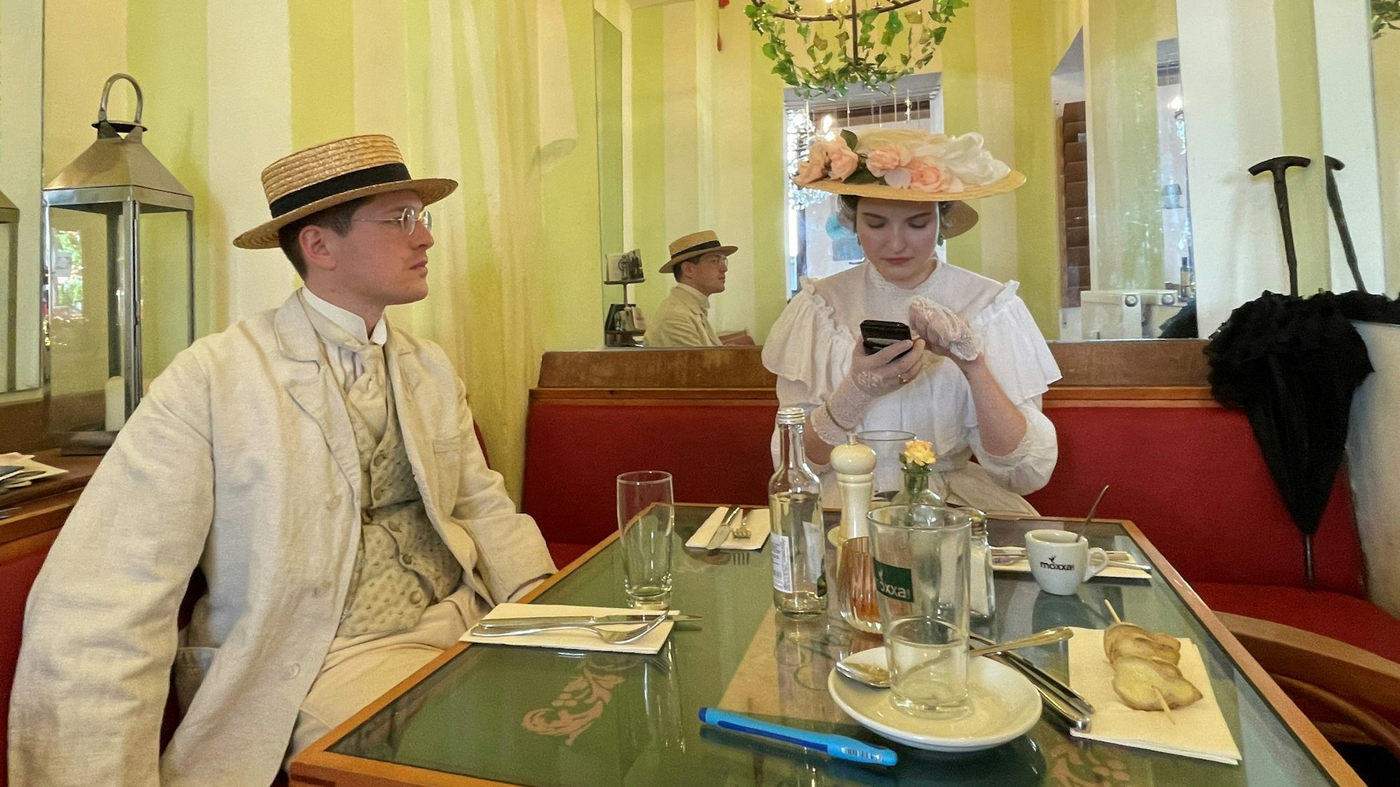 Ein Mann mit Hut und hellem Anzug mit Weste und Krawatte und eine Frau mit einem mit Blumen besetztem Hut auf dem Kopf und einem rüschenbesetzten weißen Kleid sitzen an einem Tisch in einem Café.