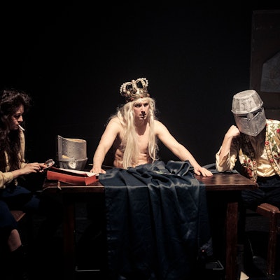 König Ubu sitzt mit freiem Oberkörper und pompöser Krone an einem Tisch, rechts und links neben ihm seine zwei Gefolgsleute mit Zigarette und Ritterhelm.